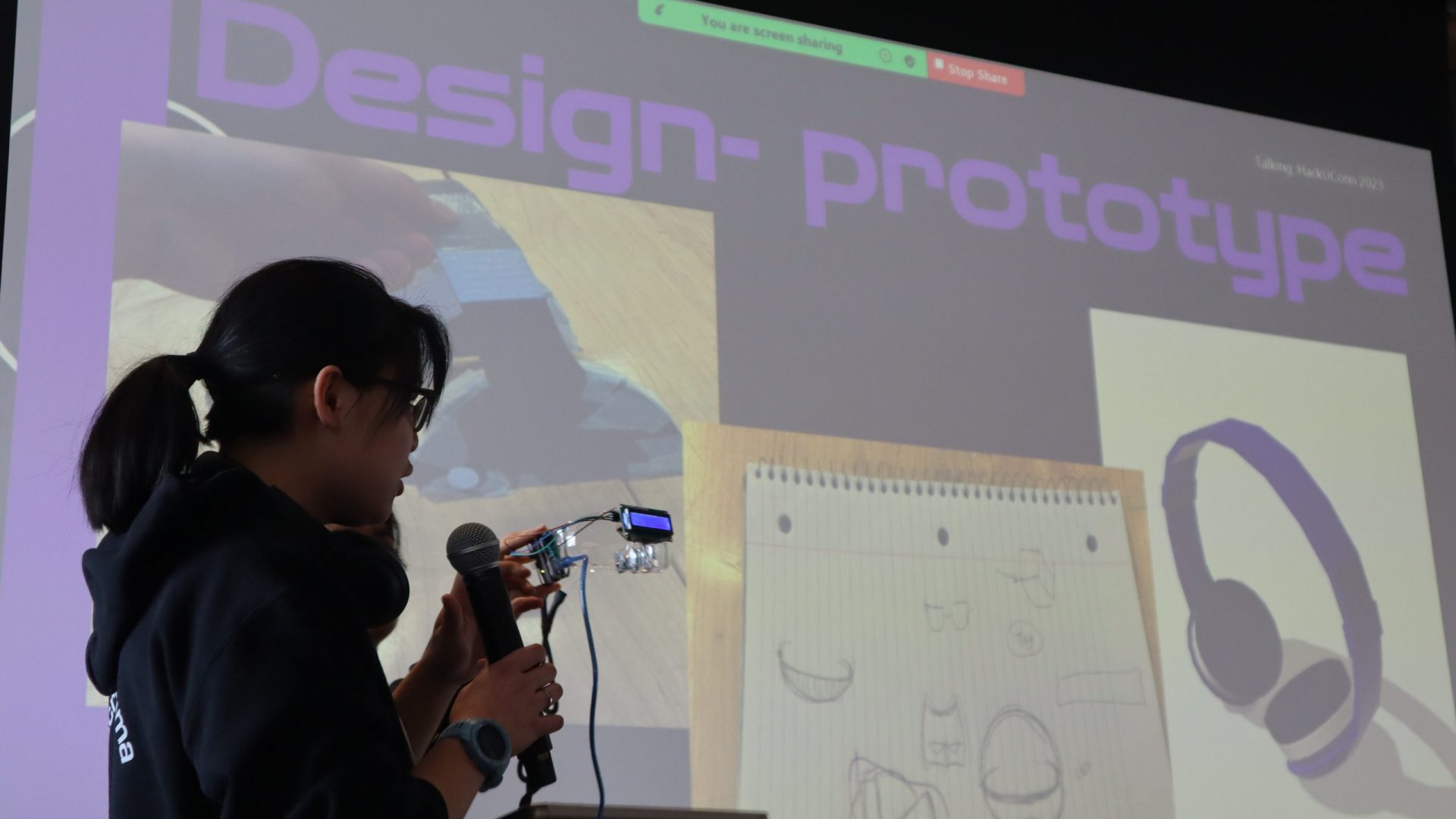 A student stands before the audience of HackUConn demonstrating their innovation by wearing their prototype, a pair of "smart glasses"